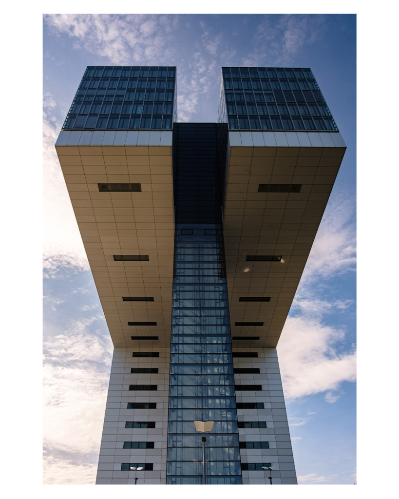 Foto im Hochformat. Ein riesiges Gebäude, ein Hochhaus, welches ab einer bestimmten Höhe nach vorne gebaut ist. Also ein Teil des Hauses steht in einem 90 Grad Winkel nach vorne. Der Teil, der nach vorne steht ist mittig noch mal geteilt und vorne nicht abgestützt. Das Gebäude ragt mächtig gegen den leicht bewölkten Himmel. 