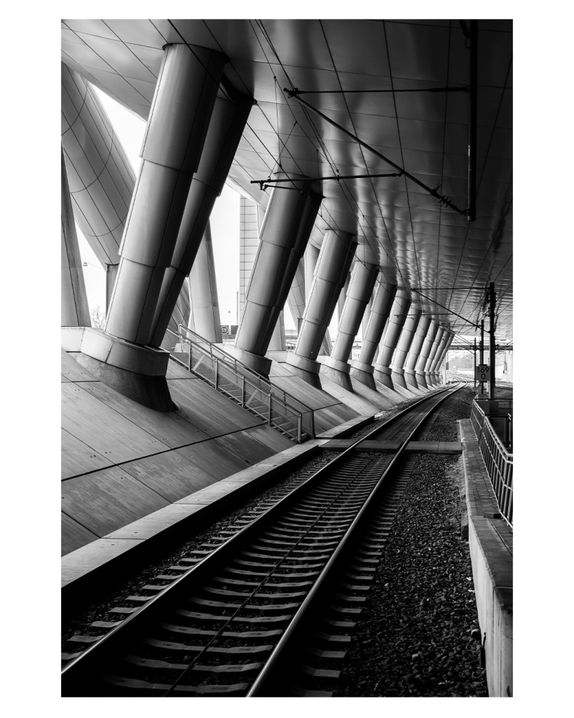 Schwarz-weiß Foto im Hochformat. Gleise an einem Bahnhof verlaufen von links unten nach rechts Mitte. Die Gleise sind überdacht, links ist eine Schräge auf der jeweils zwei dicke Säulen nebeneinander stehen, die das Dach tragen. 