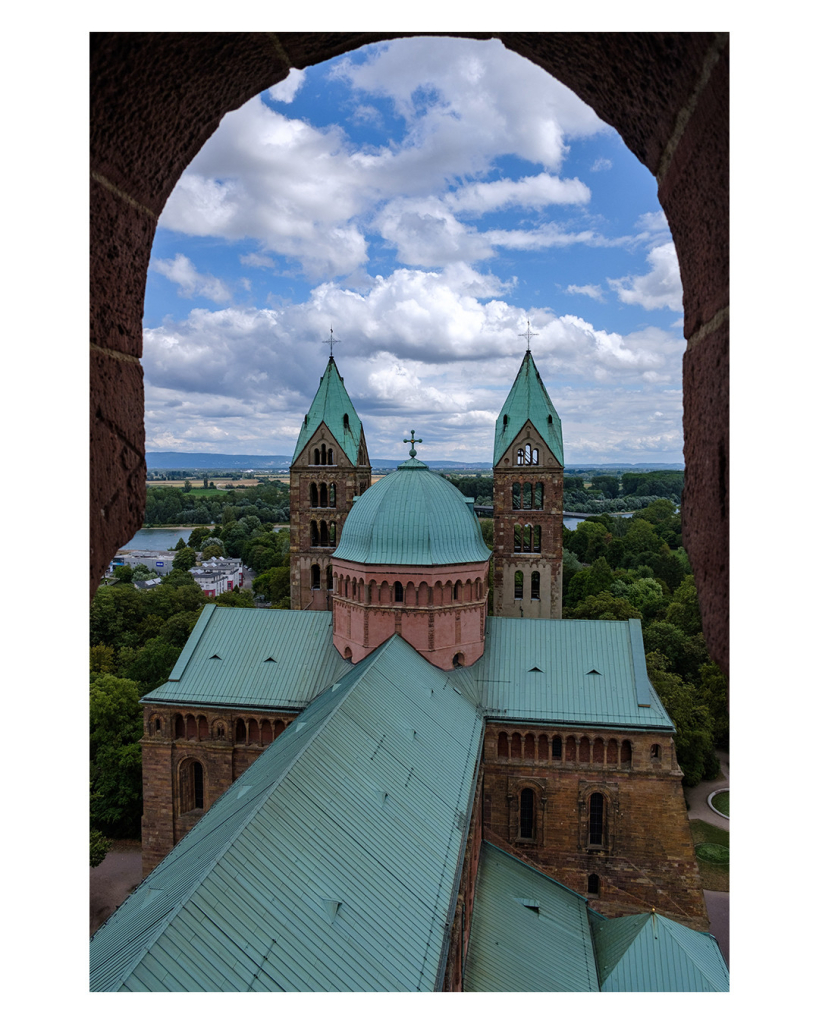 Foto im Hochformat. Blick aus einem steinernen Bogen auf das Dach des Doms in Speyer. Das Dach ist grün, weiter hinten ist eine runde Kuppel gefolgt von zwei Türmen. Der Himmel ist bewölkt. 