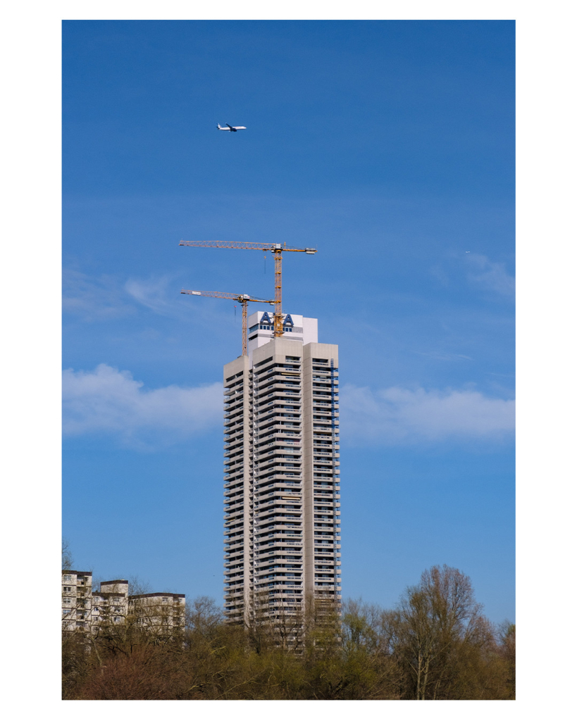 Foto im Hochformat. Ein Wolkenkratzer steht mittig im Bild. Davor sind kahle Bäume. Auf dem quadratischen Wolkenkratzer stehen zwei Kräne. Auf einem Schild steht AXA. Über dem Wolkenkratzer fliegt ein einzelnes Flugzeug. Der Himmel ist leicht bewölkt. 