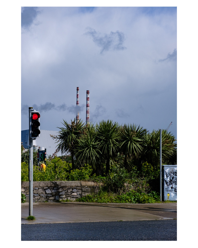 Foto im Hochformat. Blick über eine Straße. Auf der anderen Seite ist eine rote Ampel, eine niedrige Mauer und einige Palmen. Hinter den Palmen ragen in der Ferne zwei hohen rot weiß gestreifte Schornsteine empor. Vor den Schornsteinen sein einige Rauchschwaden. Der Himmel hinter und über den Schornsteinen ist bewölkt. 