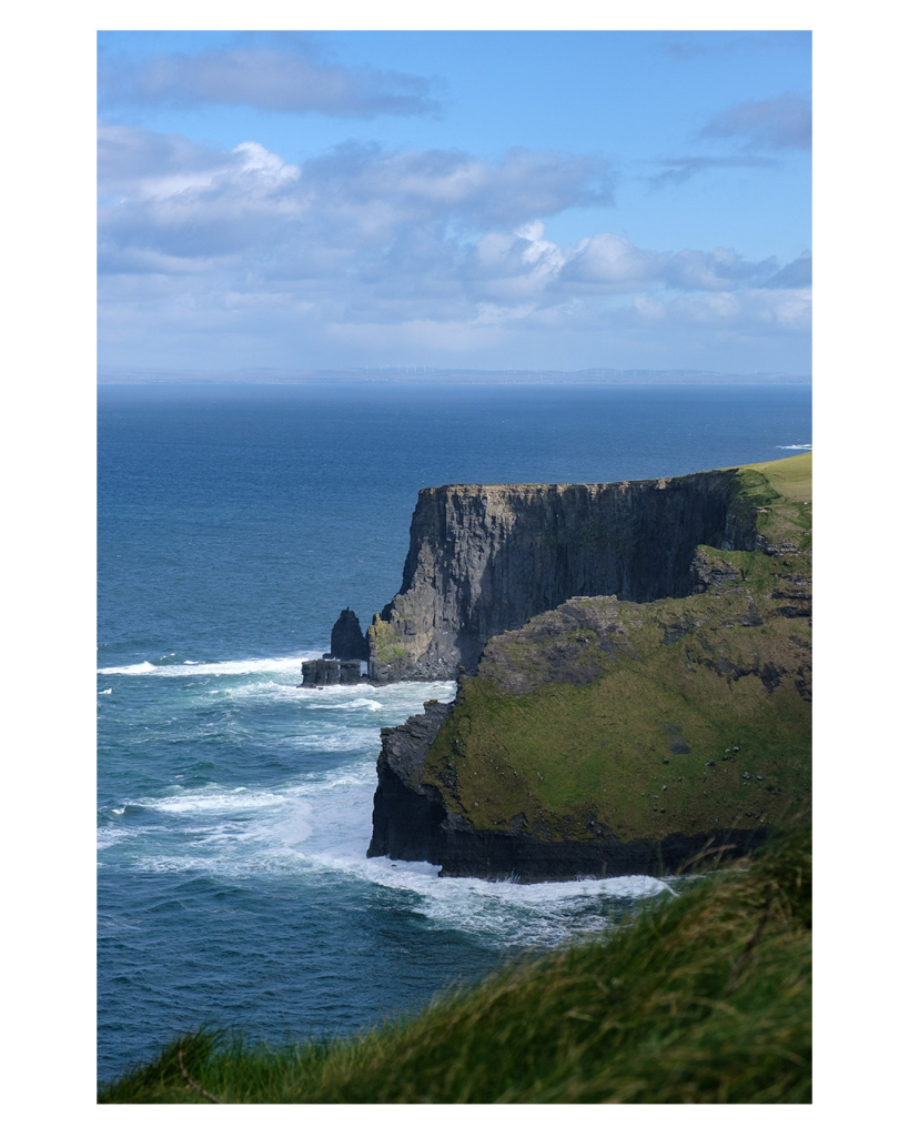 Foto im Hochformat. Blick auf die Cliffs of Moher, eine Steilküste in Irland. Zwei riesige Felswände ragen von rechts ins Bild. Das Meer unten ist weiß an den Felsen, sonst blau. Ganz im Vordergrund ist Gras, leicht unscharf. Der Himmel ist leicht bewölkt. 