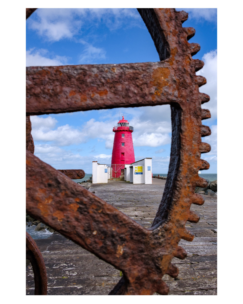 Foto im Hochformat. Ausschnitt eines verrosteten Zahnrades. Durch die Speichen sieht man einen knallroten Leuchtturm, vor dem Unterstellmöglichkeiten aus weißem Stein sind. Der Turm steht am Ende eines steinernen Piers. Der Himmel ist leicht bewölkt. 