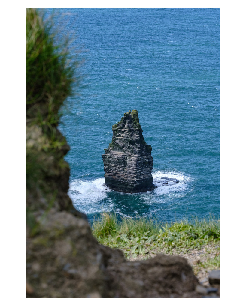 Foto im Hochformat. Teleaufnahme eines großen Felsen im Meer. Das Meer ist blau, um den Felsen herum ist weiße Gischt. Im Vordergrund ist unscharf links und unten ein Felsen angeschnitten. Davor ist Gras. 