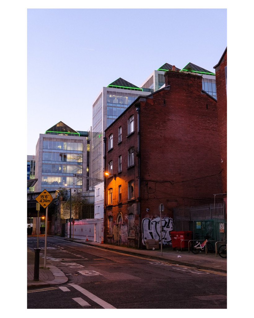 Foto im Hochformat. Blick einer Straße in einer Großstadt entlang. Rechts ist ein altes Gebäude aus roten Backsteinen. Es ist unten mit Graffiti voll gesprüht. Zur Straße hin ist am Gebäude eine Straßenlaterne angebracht, die einen Teil des Hauses gelblich rot beleuchtet. Hinter dem Gebäude sind mehrere große moderne Finanzgebäude, mit viel Glas und spitzen Dächern, die Grünen leuchtende Streifen haben. Der Himmel ist klar, es ist Abend. 