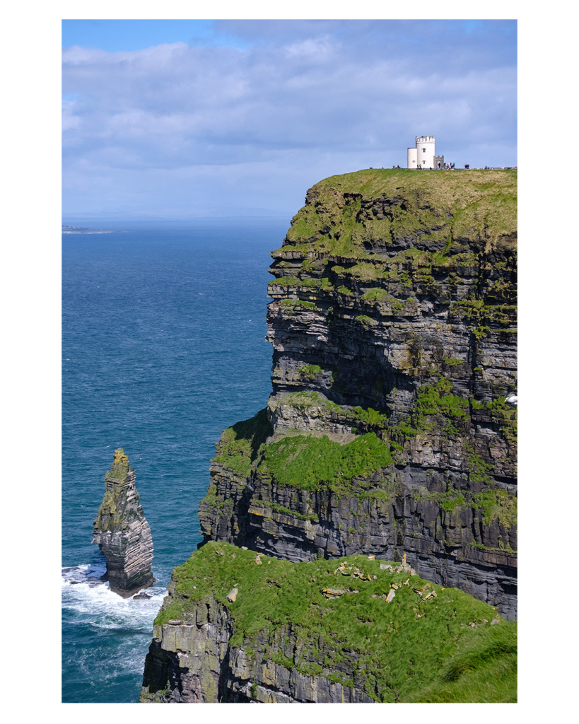 Foto im Hochformat. Blick auf eine riesige Klippe, die von rechts ins Bild ragt. Die felsige Klippe ist an den flacheren Stellen mit Gras bewachsen. Oben auf der Klippe ist ein weißes rundes Gebäude. Im Gegensatz zur Klippe sieht dieses sehr winzig aus. Vorname Gebäude laufen Menschen umher, die entsprechend noch winziger aussehen. Unten im Meer ist noch ein einzelner Felsen der aus dem Meer ragt. Der Himmel ist bewölkt. 