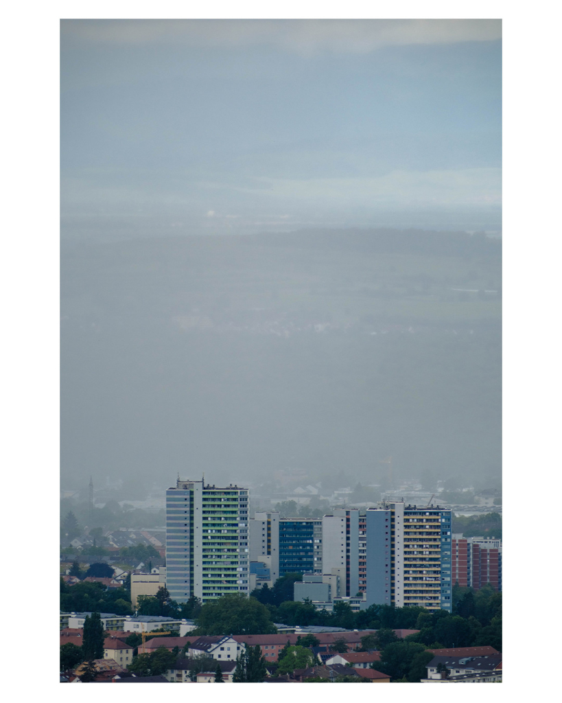 Foto im Hochformat. Teleaufnahme mehrere Hochhäuser in einem Wohngebiet. Die Hochhäuser machen das untere Drittel des Bildes aus, darüber geht gerade Regen nieder, durch den Berge und der Himmel dahinter kaum zu erkennen sind. 