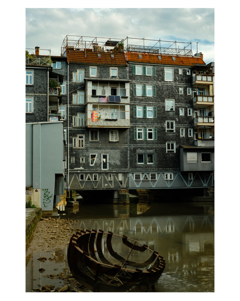 Foto im Hochformat. Blick auf ein altes blau-graues Gebäude vertäfelter Fassade. Am Haus sind ein paar Balkone, die Fenster haben grüne Fensterläden, das Dach besteht aus roten Ziegeln und es gibt Dachterassen mit Geländer. Unter dem Gebäude fließt Wasser durch, welches im Vordergrund des Bildes ist. Ganz vorne liegt das Gerippe eines Ruderbootes. Der Himmel ist bewölkt. 