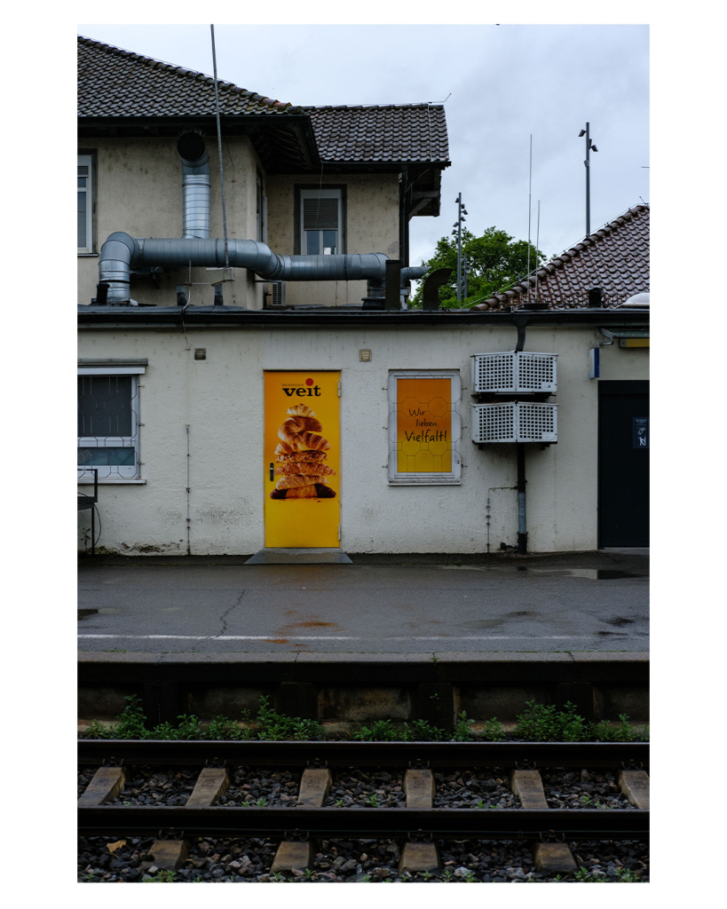 Foto im Hochformat. Blick über Bahngleise auf ein Gebäude. Das Gebäude ist älter, hell verputzt und daran befestigt sind Klimaanlage und dicke Rohre. An einer Tür und an einem Fenster ist gelbfarbene Werbung für die Bäckerei Veit. Darauf abgebildet sind Backwaren und der Spruch „Wir lieben Vielfalt“. Das gelb sticht aus dem sonst tristen Anblick stark hervor. 