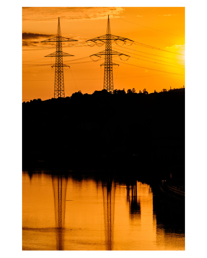 Foto im Hochformat. Sonnenuntergang. Links oben im Bild sind zwei Hochspannungsmasten, von denen links und rechts zahlreiche Kabel weggehen. Davor sind Büsche und Bäume, die aber komplett im Schatten versinken. Der Himmel ist gelblich orange. Unten im Bild ist ein Fluss, in dem sich alles in leicht gekräuseltem Wasser spiegelt. Rechts oben ist knapp die untergehende Sonne noch im Bild.   