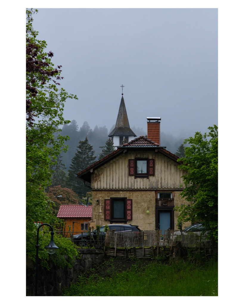 Foto im Hochformat. Ein altes einzelnes Wohnhaus mit hölzerner Vertäfelung und Spitzdach. Es ist eingerahmt von Büschen und Bäumen, dahinter ragt der Turm einer Kirche hervor. Es regnet und der Himmel ist mit Nebel verhangen. 