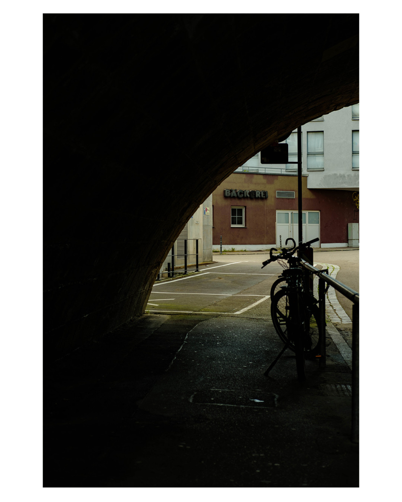 Foto im Hochformat. Blick von unter einer Brücke auf eine Straße. Von links unten nach rechts oben ist der Bogen der Brücke. Dieser versinkt komplett im Schatten. Rechts ist ein Geländer an dem Fahrräder angeschlossen sind. Der Blick geht nach vorne auf eine braune Hauswand auf der mit großen schwarzen Buchstaben Bäckerei steht. Unter der Schrift ist ein kleines Fenster. Bis auf die Schrift deutet nichts auf ein Ladengeschäft hin. 