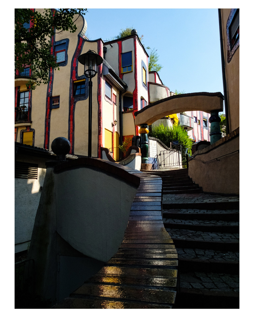 Foto im Hochformat. Blick von unten auf ein Hundertwasserhaus. Der Blick wird geführt von einer Treppe bzw. von einer Mauer, die als Geländer für die Treppe dient. Das Haus ist hell verputzt mit vielen bunten Verzierungen und Schnörkeln. 