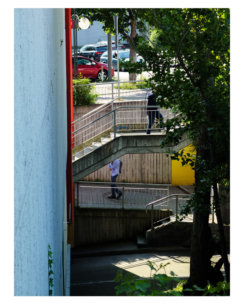 Foto im Hochformat. Blick einer Hauswand entlang auf mehrere Treppen und schräge Gehwege. Zwei Menschen laufen gerade nach oben, bei beiden ist der Kopf durch eine Mauer bzw. einen Baum abgeschnitten und nicht zu erkennen. 