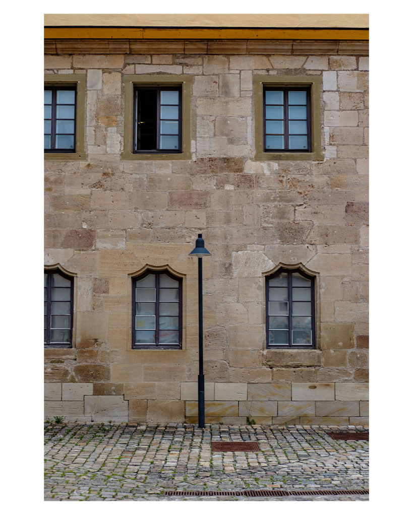 Foto im Hochformat. Blick auf die Hauswand eines alten Gebäudes. Vier rechteckig Fenster sind komplett im Bild, zwei weitere sind links jeweils in der Mitte vom Bildrand geteilt. Die Fenster sind ebenfalls alt, die unteren haben jeweils oben eine geschwungene Form. Mittig im Bild, aber nicht mittig zwischen den Fenstern steht eine einzelne Straßenlaterne. Der Boden vorm Haus ist Kopfsteinpflaster. 