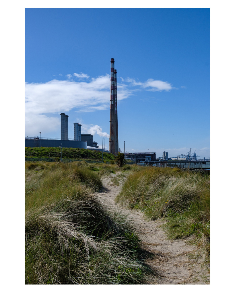 Foto im Hochformat. Blick über mit Gras bewachsenen Dünen auf zwei hohe Industrieschornsteine, die weiß rot gestreift sind. Durch die Dünen führt ein sandiger Pfad genau Richtung Schornsteine. Neben den Schornsteinen sind kleinere Industriegebäude. Rechts in etwas Entfernung ist ein industrielle Hafenanlage. Der Himmel ist blau, nur hinter und links von den Schornsteinen ist eine große weiße Wolke. 