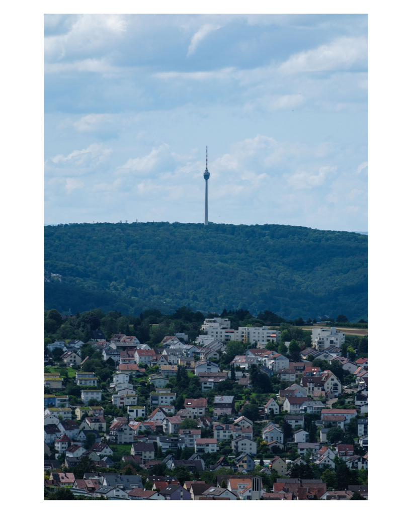 Foto im Hochformat. Teleaufnahme. Im unteren Teil des Bildes ist ein Wohngebiet, darüber ein bewaldeter Berg und auf dem Berg, mittig im Bild steht ein Fernsehturm. Durch den zoom sieht alles viel näher beieinander aus, als es in Wirklichkeit ist. Der Himmel ist leicht bewölkt. 