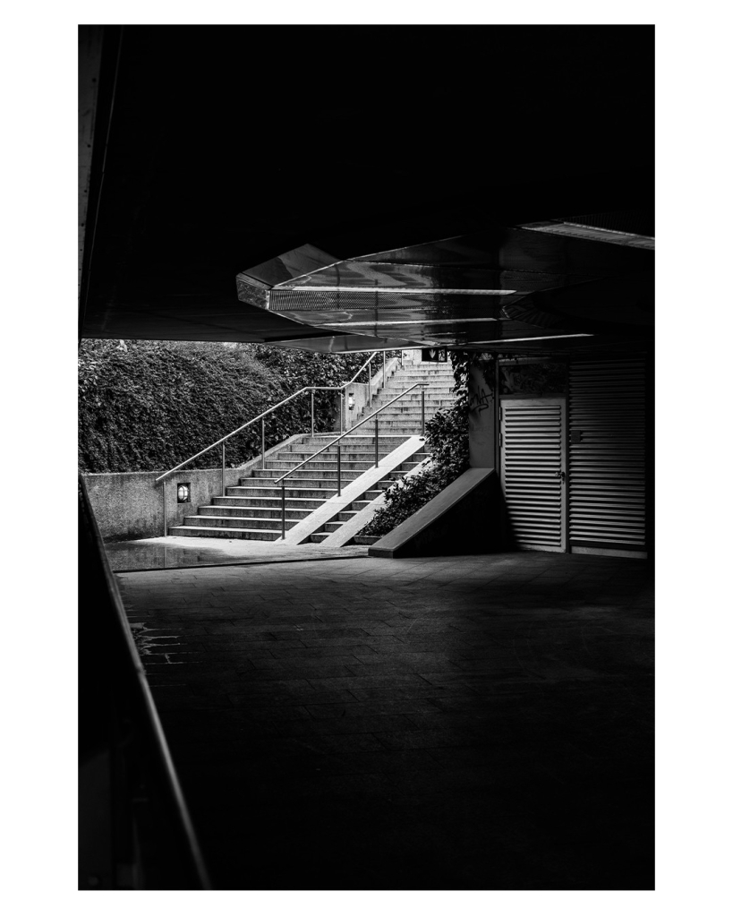 Schwarz-weiß Foto im Hochformat. Blick aus einer Unterführung Richtung einer Treppe, die nach oben führt. Alles in der Unterführung versinkt im Schatten. Die Treppe wird hell von der Sonne angestrahlt. An der Decke der Unterführung ist eine runde Konstruktion. Links ist ein Geländer, welches Richtung Treppe führt. 