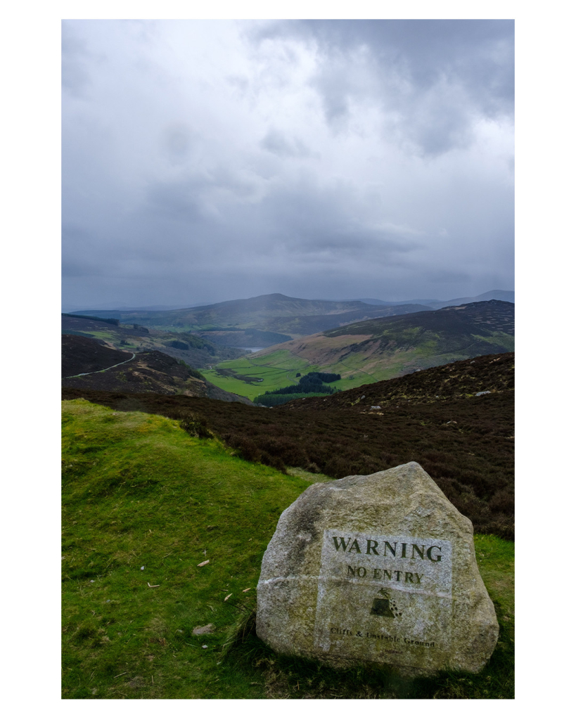 Foto im Hochformat. Blick über eine hügelige Landschaft, die aus Wiesen, Wäldern und Torf besteht. Im Vordergrund ist ein Felsen, auf diesem steht „Warning. No Entry. Cliffs & unstable Ground“. Dazu ein Piktogramm einer Person, die von einem Kliff fällt. Es ist stark bewölkt und regnerisch. Weiter hinten fallen Sonnenstrahlen durch die Wolken. 