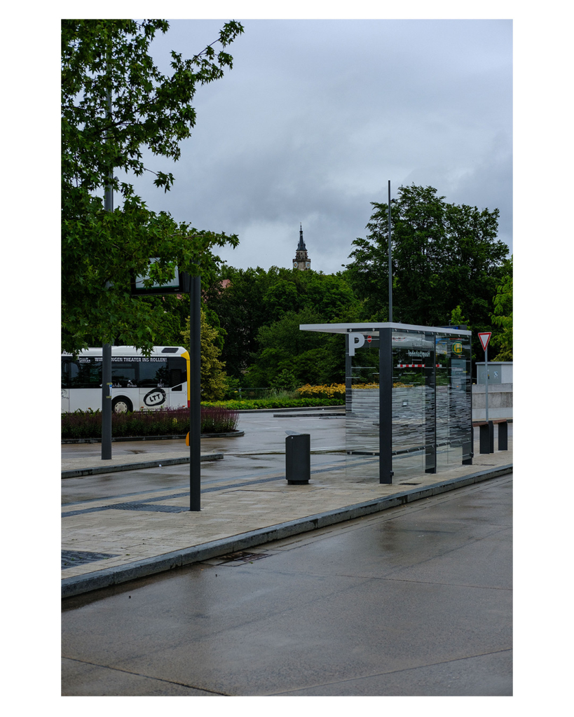 Foto im Hochformat. Blick auf einen fast leeren Busbahnhof. Eine Bushaltestelle verläuft diagonal durchs Bild, an ihr ist gläsernes Dach mit gläsernen Wänden an drei Seiten um sich unterzustellen. Auf den Wänden sind viele horizontale Streifen als Muster. Außerdem ein großes P. Links im Bild an einer anderen Haltestelle steht ein weißer Omnibus. Zwischen Bus und dem gläsernen Dach ist ein Mülleimer. Hinter dem Busbahnhof sind viele grüne Bäume, darüber ragt die spitze eines Kirchturmes hervor. Diese ist genau über dem Mülleimer. Der Himmel ist bewölkt und es hat geregnet. 