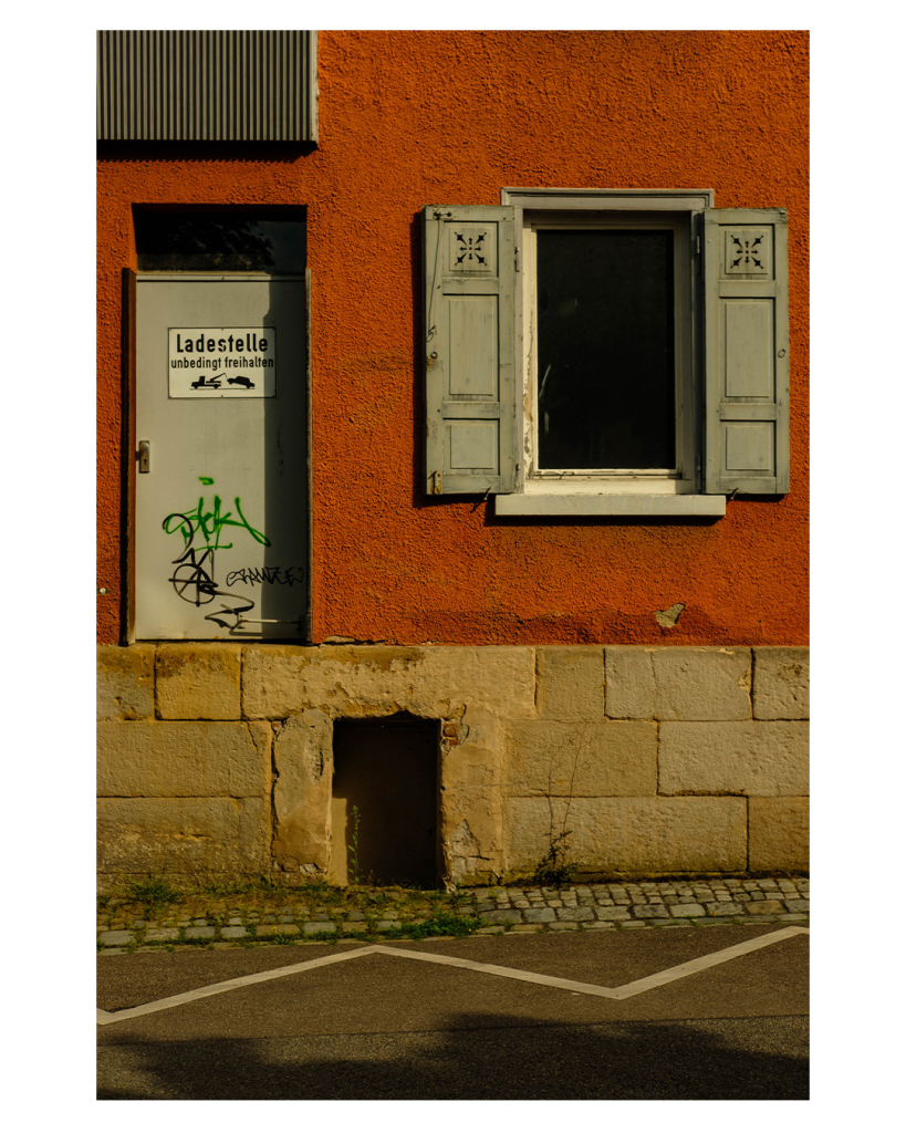 Foto im Hochformat. Blick auf eine Hauswand. Unten ist ein Streifen helle Mauer, darin ist auf Bodenhöhe ein Fenster. Darüber ist rötlicher Putz, darin ist eine Tür mit einem Schild auf dem Ladestelle unbedingt freihalten steht. Daneben ist ein Fenster mit zwei geöffneten Fensterläden. Die Fensterläden sind grau, so wie die Tiere. Alles zusammen sieht aus wie ein Emoji mit aufgerissenem Mund. 