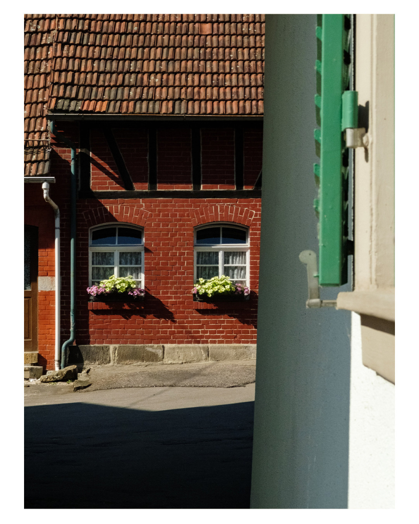 Foto im Hochformat. Blick an einer weißen Hauswand entlang auf einen Hinterhof. An der weißen Hauswand ist Teil eines Fensters mit grünen Fensterläden zu sehen. Am Ende des Hofes ist ein Gebäude aus roten Backsteinen. Zwei Fenster mit weißen Fensterrahmen und Blumentöpfen an der Fensterbank. Direkt vorm Haus fällt die Sonne hin, dahinter ist dunkler Schatten. Die weiße Hauswand liegt ebenfalls halb im Schatten. 