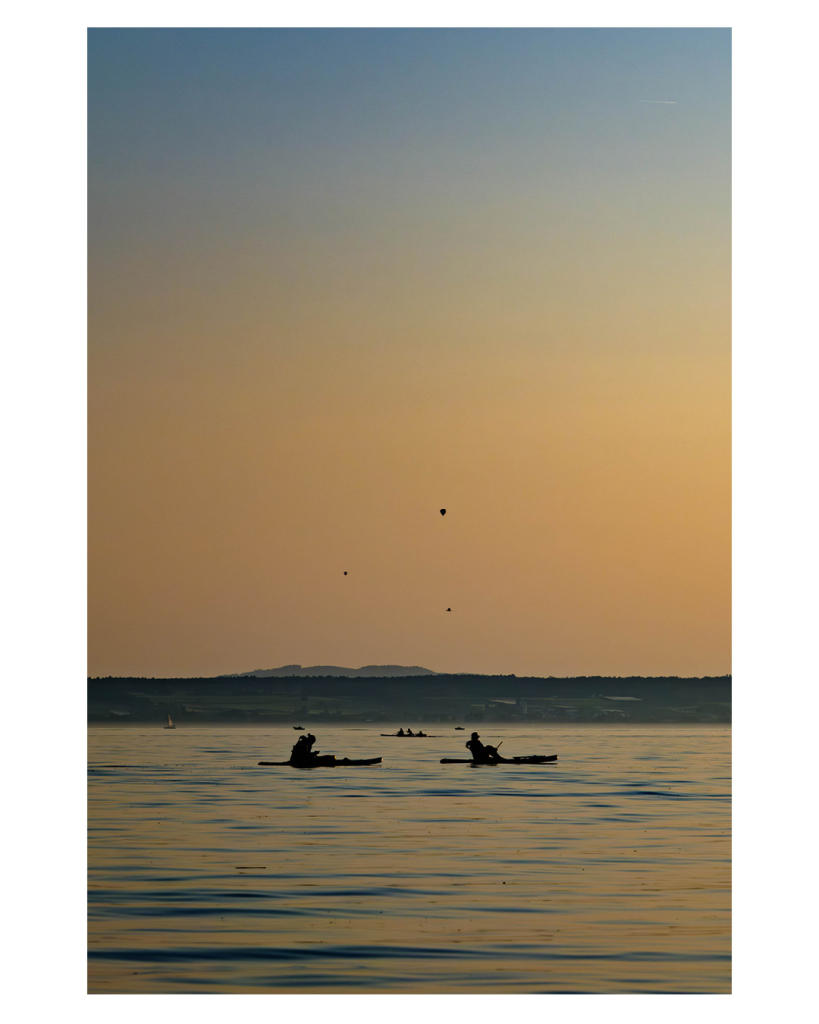 Foto im Hochformat. Blick auf einen See kurz vor Sonnenuntergang. In einiger Entfernung paddeln zwei Menschen jeweils auf einer Art  Surfboard. Sie sind nur als Silhouette wahrzunehmen. Genau zwischen ihnen in noch weiterer Entfernung ist ein weiteres flaches Boot mit drei Insassen. Am Horizont ist das gegenüberliegende Ufer, ein flacher dunkler Streifen im Bild. Darüber, über die Hälfte des gesamten Fotos ist der Wolkensteins Himmel. Erst gelblich-orange, weiter oben dann hellblau. Am Himmel sind genau über den beiden Paddlern drei Punkte, die ein gleichseitiges Dreieck ergeben. Es sind zwei Vögel und ein Heißluftballon. Dies erkennt man aber nur, indem man rein zoomt ins Bild. 