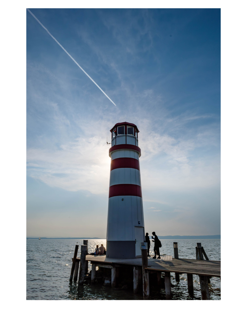 Foto im Hochformat. Auf einem Holzsteg steht ein relativ kleiner Leuchtturm. Er ist weiß mit zwei mit drei roten Streifen und rotem Dach. Oben sind rundherum Fenster. Die Sonne ist genau hinter dem Turm und bestrahlt diesen. Neben dem Turm sind einige Menschen, hinter dem Turm Wasser bis zum Horizont. Aus der linken oberen Ecke kommt der Kondensstreifen eines Flugzeuges, welches genau über dem Turm ist. Der Himmel ist leicht bewölkt. 