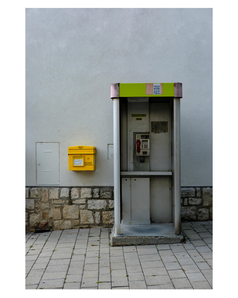 Foto im Hochformat. Eine österreichische Telefonzelle an einer Hauswand. Links daneben hängt ein gelber Briefkasten an der Wand. Die Telefonzelle hat keine Türe, oben ist sie grün, an den Ecken rosa. Auf einem Schild steht Telekom Austria. Die Wand ist weiß, unten ist ein Streifen aus Steinfliesen. Da der Gehweg leicht bergab geht, steht die Telefonzelle auf einem Podest aus Beton, welches die Schräge ausgleicht. Neben dem Briefkasten ist eine weiße Klappe in der weißen Wand auf dem ein Blitz Symbol ist. 