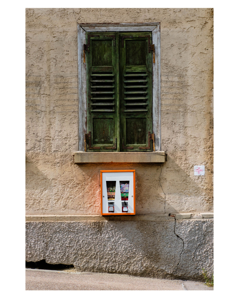 Foto im Hochformat. Eine alte Hauswand mit bröckligem dreckigem gelblichen Putz. Mittig ist ein Fenster mit geschlossenem grünen Fensterladen. Im Fensterladen sind Schlitze, Abdrücke dieser Schlitze sieht man rechts und links neben dem Fenster. Genau mittig unter dem Fenster ist ein weißer Kaugummiautomat mit orangefarbenem Rand. Wiederum darunter ist an der Wand ein grauer Streifen der bis zum Boden geht. Der Fußweg vor dem Haus geht leicht schräg bergab.  