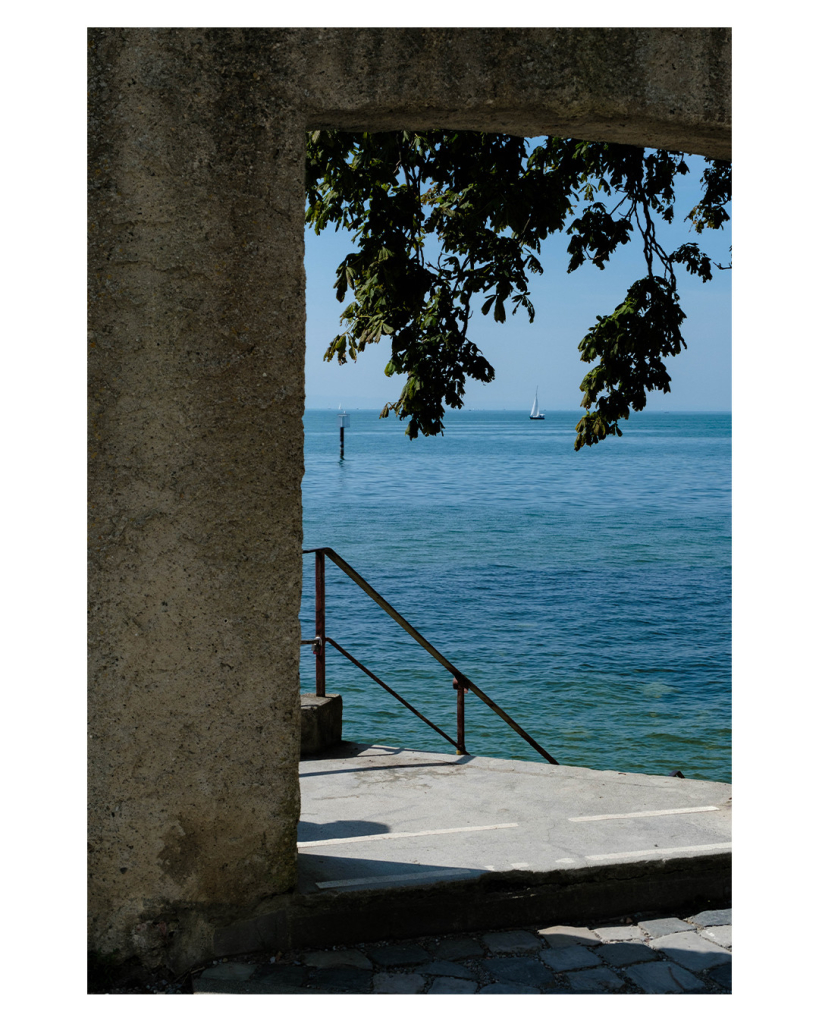 Foto im Hochformat. Blick durch einen Durchgang in einer Mauer auf den Bodensee. Der Durchgang ist nur links und oben zu sehen und er ist im Schatten. Dahinter, in der Sonne, führt ein Treppengeländer nach unten. Von oben ragen Äste mit Blättern, genau zwischen zweien ist eine Segelschiff am Horizont auf dem See. Der Himmel ist klar. 