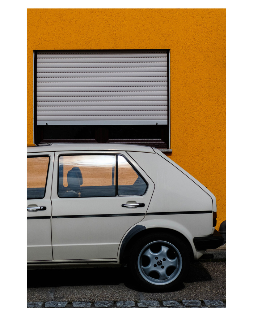 Foto im Hochformat. Ein grauer Volkswagen Golf I (vermutlich) steht seitlich vor einer knallorangenen Hauswand an der ein Fenster mit fast geschlossenem grauem Rollladen. Vom Auto ist nur der hintere Teil im Bild. 