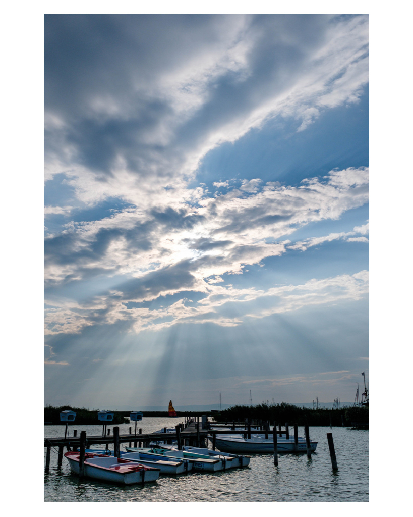 Foto im Hochformat. Blick auf eine Bucht in einem See, vorne im Bild ist ein Steg mit kleinen Booten. Weiter hinten ist ein kleines Segelboot auf dem Wasser. Der Himmel ist leicht bewölkt, hinter den Wolken ist die Sonne, deren Strahlen deutlich zu sehen sind. Sie gehen bis zum See hinab. 