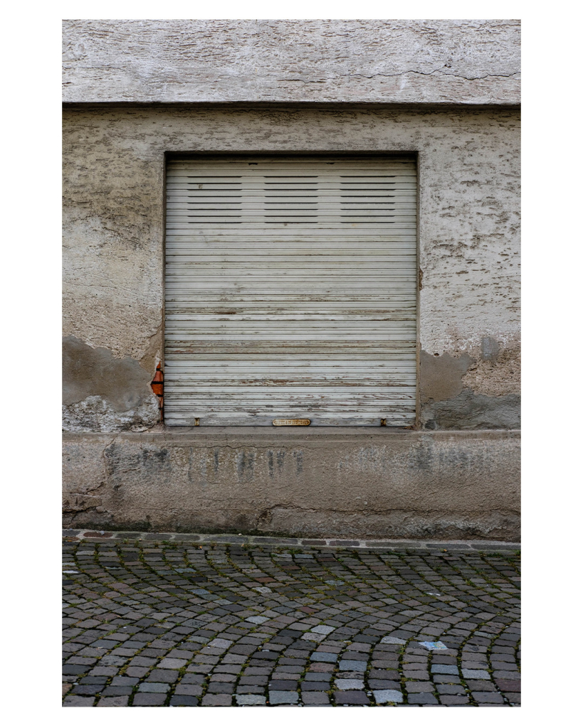 Foto im Hochformat. Eine graue Wand, in der ein Fenster ist. Der abgeranzte graue Rollladen ist unten. Über dem Fenster ist ein grauer Absatz. Vor der Wand ist dunkelgrauer Kopfsteinpflaster. Alles sieht unfassbar eintönig, alt und kaputt aus. Auf der linken Seite des Fensters ist der Putz aufgeplattt, knallroter Backstein schaut heraus. 