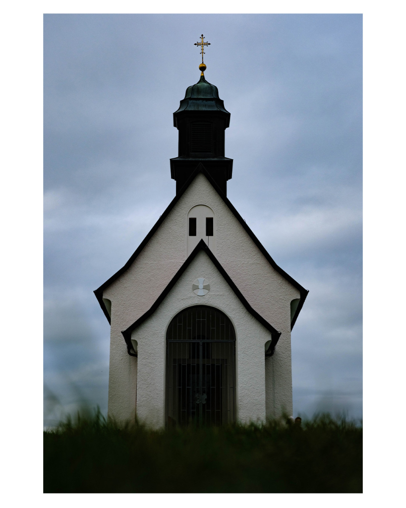 Foto im Hochformat. Blick einen Hügel empor auf eine weiße Kapelle mit dunklem Dach. Die Kapelle hat einen kleinen Vorbau mit Dach, dahinter ist da eigentliche Gebäude. Oben ist ein kleiner Turm mit einem Kreuz darauf. Im Vordergrund ist Gras, es ist unscharf und dunkel. Der Himmel ist bewölkt. Das ganze Bild sieht recht düster aus. 