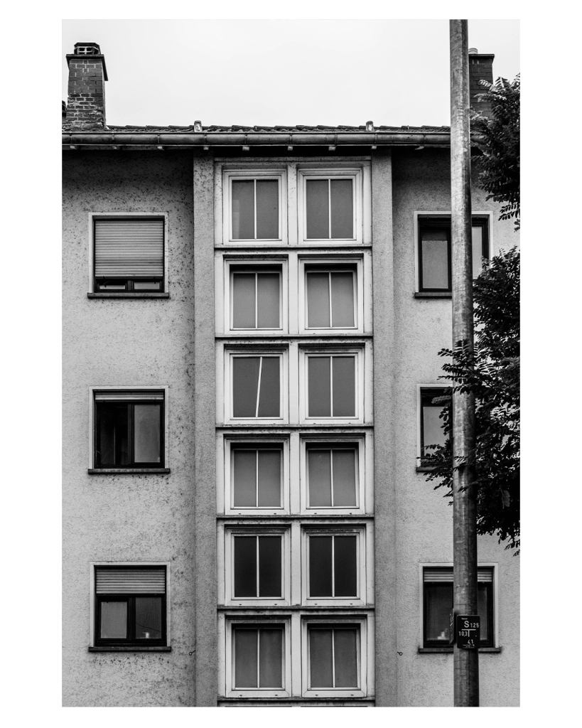 Schwarz-weiß Foto im Hochformat. Gerader Blick auf ein altes Wohnhaus. Mittig sind sechs Doppelfenster mit weißem Fensterrahmen übereinander. Rechts und links davon sind jeweils drei Fenster. Alle sind unterschiedlich geöffnet bzw. haben unterschiedlich geschlossene Jalousien. Direkt vor den rechten Fenstern ist ein Laternenpfahl. Die Fensterrahmen sind alt und aus Holz und einer ist schief und fällt direkt ins Auge. Alles im Bild ist sehr symmetrisch. 