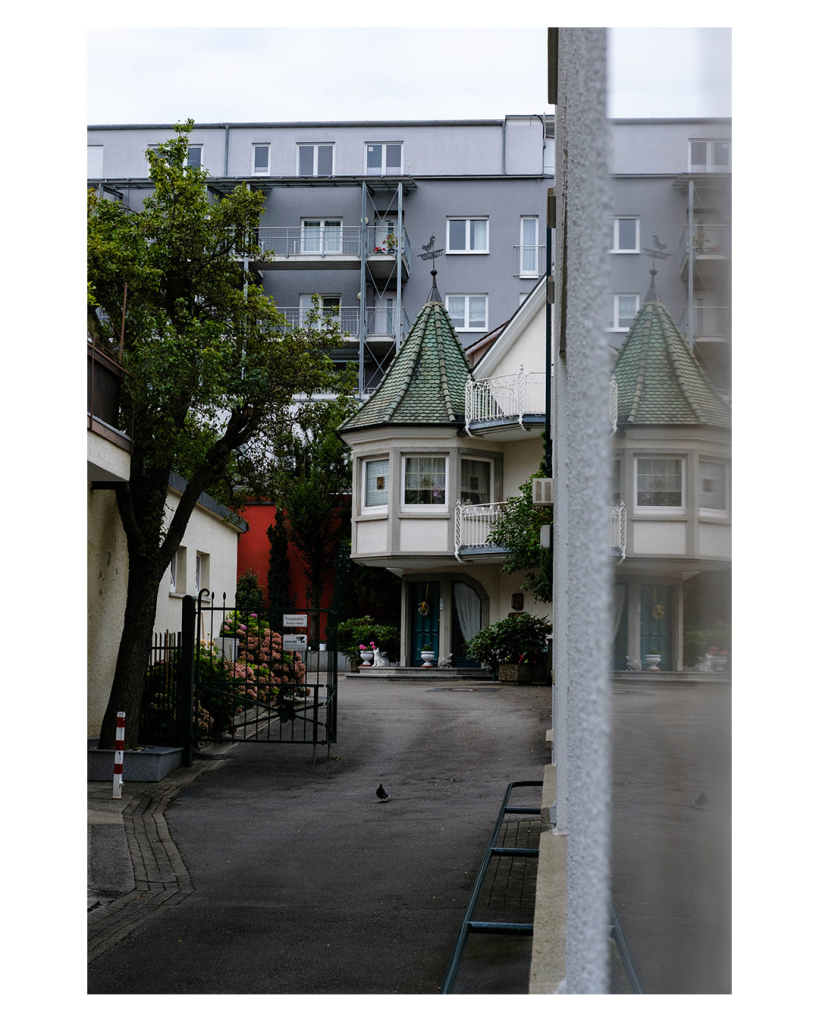 Foto im Hochformat. Blick entlang einer Einfahrt in einer Stadt. Am Ende der Einfahrt ist ein altes Gebäude mit einem eckigem Turm mit grünem Spitzdach. Auf der Spitze ist ein Wetterhahn. Hinter dem Gebäude ist belangloses graues mehrstöckiges Wohngebäude. Vor dem alten Hause sind Blumen und ein Gittertor. Rechts neben der Kamera ist eine Wand mit reflektierender Fläche. Dort spiegeln sich Teile der Gebäude wieder. 