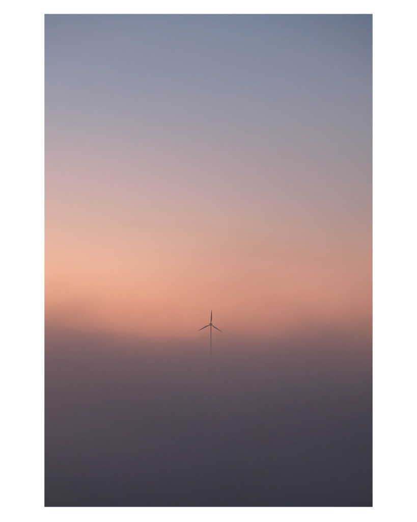 Foto im Hochformat. Sonnenaufgang, alles ist neblig. Darüber ist der Himmel, am Horizont rötlich, darüber hellblau. Das einige, was im Nebel zu sehen ist, ist der obere Teil einer Windkraftanlage. Einer der drei Rotoren des Propellers zeigt genau nach oben. 