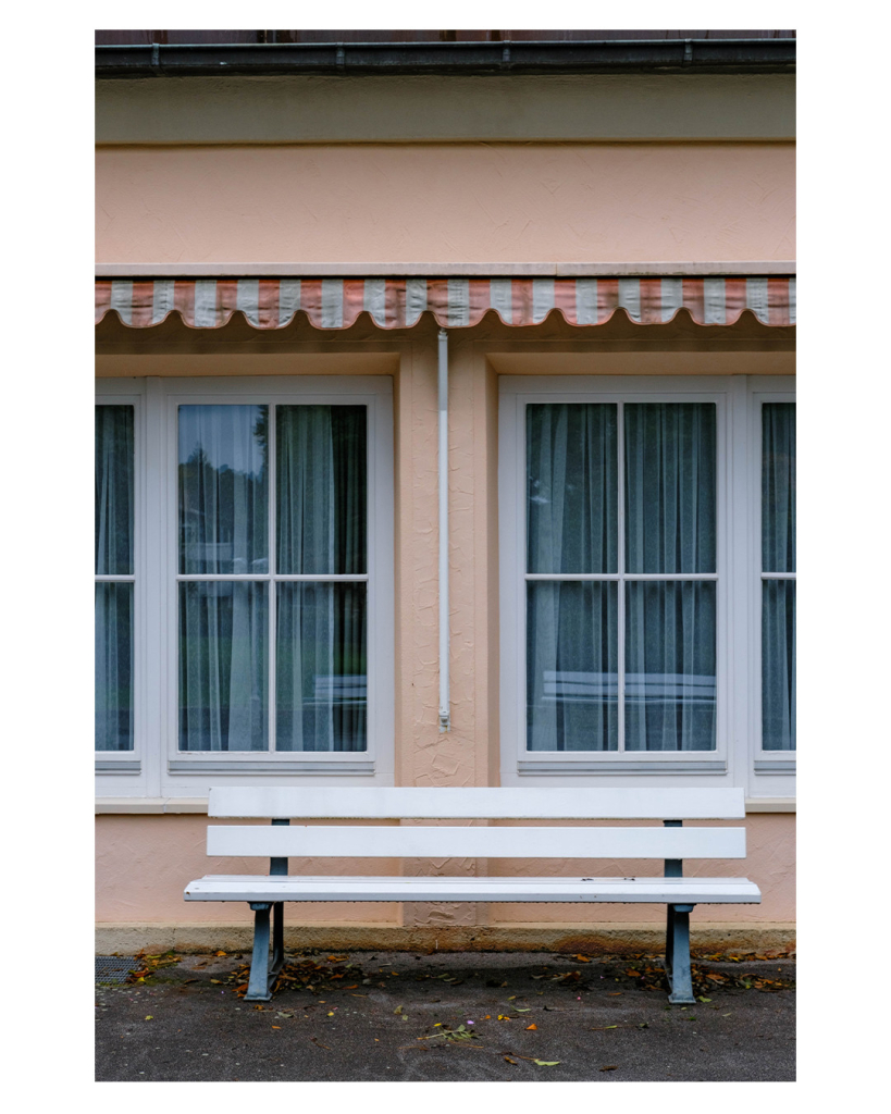 Foto im Hochformat. Blick auf eine pastellfarbene Hauswand. Zwei rechteckige Fenster mit weißen Rahmen. Hinter den Fenstern sind weiße Vorhänge. Zwischen den Fenstern ist die weiße Stange einer Markise, welche eingefahren über den Fenstern entlang geht. Die Markise ist orange weiß gestreift. Vor der Wand steht eine weiße Parkbank. Alles im Bild ist sehr symmetrisch angeordnet.  