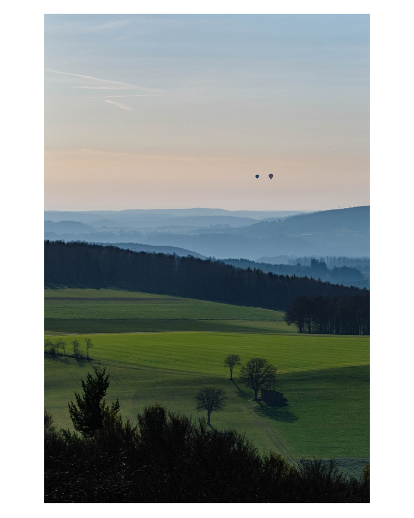 Foto im Hochformat. Blick über die schwäbische Alb von einem hohen Punkt aus. Im Vordergrund sind Wiesen mit vereinzelten Bauern, dann kommen Wälder. Dahinter Berge und Täler in denen der Nebel hängt. Am Himmel sind zwei Heißluftballons relativ nah beieinander. Der Himmel ist blau mit wenigen Wolken. 