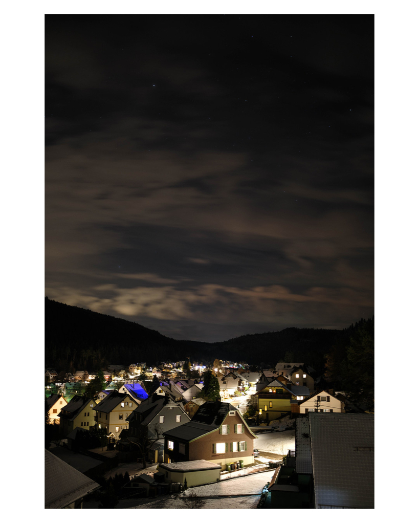 Foto im Hochformat. Es ist nacht, Blick über die beleuchteten Häuser eines verschneiten Dorfes. Hinter den Häusern sind links und rechts Berge, die allerdings nur als Silhouette zu erkennen sind. Darüber ist der bewölkte Nachthimmel mit vielen Wolken und einigen wenigen Sternen.  