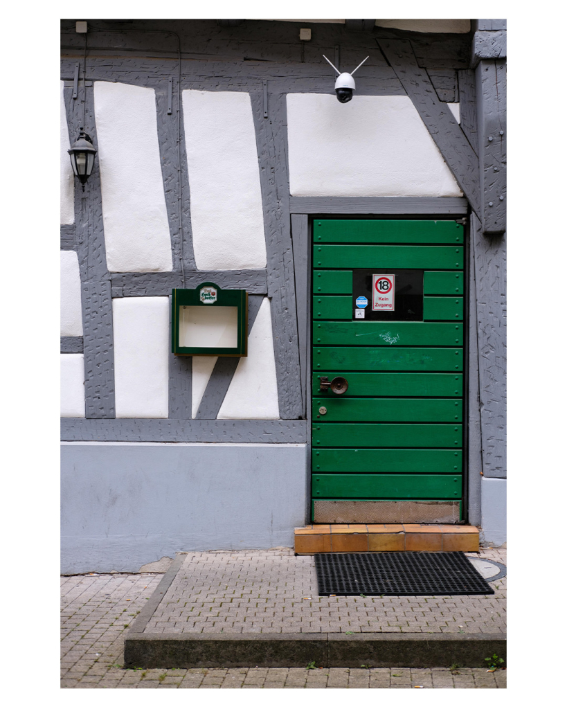 Foto im Hochformat. Ausschnitt einer Wand eines Fachwerkhauses. Weißer Stein, graue Balken. Darin ist eine Tür aus grünen Brettern. Oben ist ein Fenster, darin hängt ein Schild, eine 18 in einem roten Kreis, kein Zugang. Neben der Türe ist ein Kasten, in dem normalerweise die Speisekarte eines Restaurants hängt. Der Kasten ist aber leer. Über der Türe ist eine Überwachungskamera. Vor der Türe ist eine quadratische ca. 10 cm hohe Plattform aus Knochensteinen. 