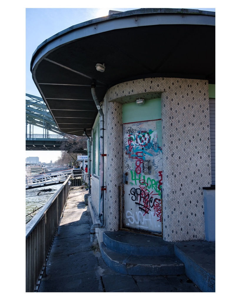 Foto im Hochformat. Rechts ein Gebäude an einer Promenade. Links Geländer, dahinter Wasser. Die Wand des Gebäudes ist aus kleinen hellen Fliesen, immer wieder sind dunkle dabei. Die Ecke des Gebäudes ist abgerundet, darin ist eine grüne Tür, die vollgekritzelt mit Graffiti ist. 
