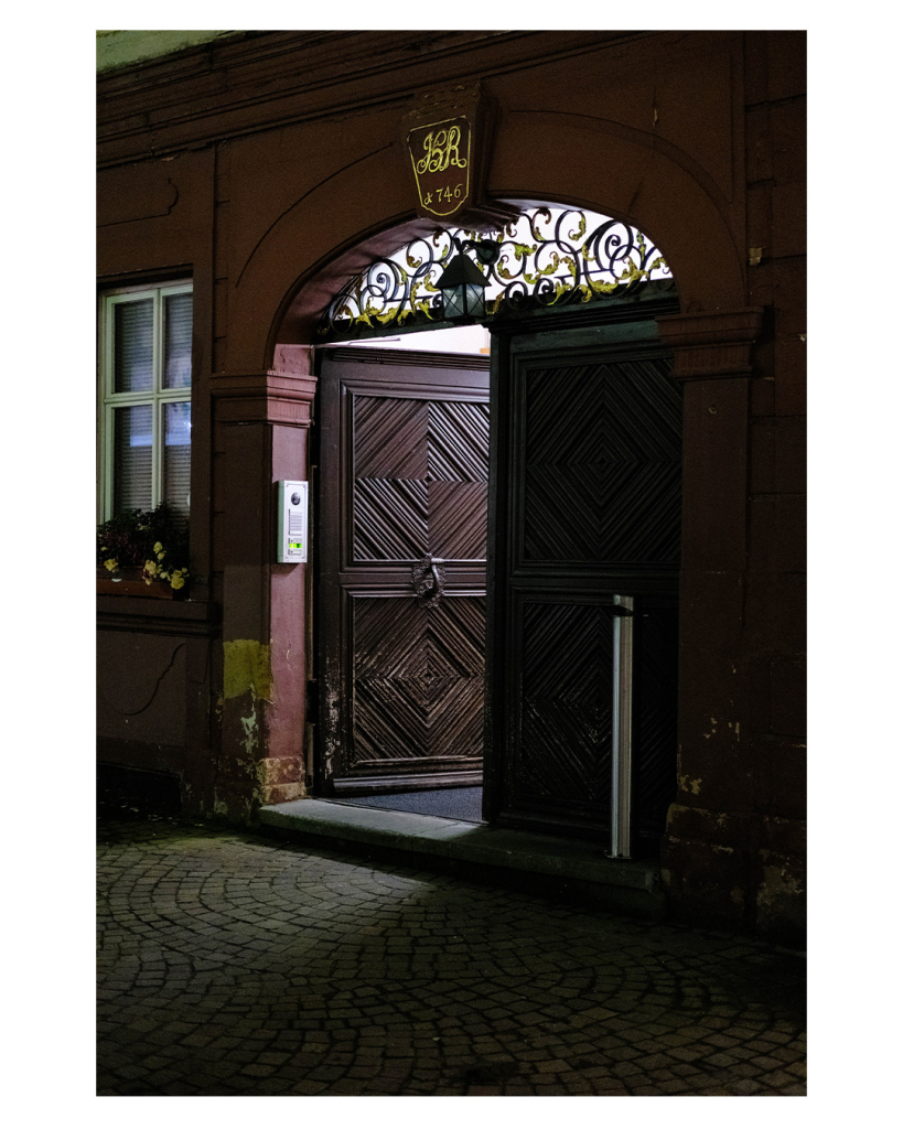 Foto im Hochformat. Nachts. Schräger Blick auf eine geöffnete Türe an einem alten Gebäude. Die Türe ist aus braunem Holz und hat ein rautenförmiges Muster. In der Mitte ist ein großer Türknauf. Es sind zwei Türen, nur die linke ist geöffnet. Über den Türen ist ein runder Bogen, darin spielförmige Verzierungen. Aus der Türe scheint Licht. 