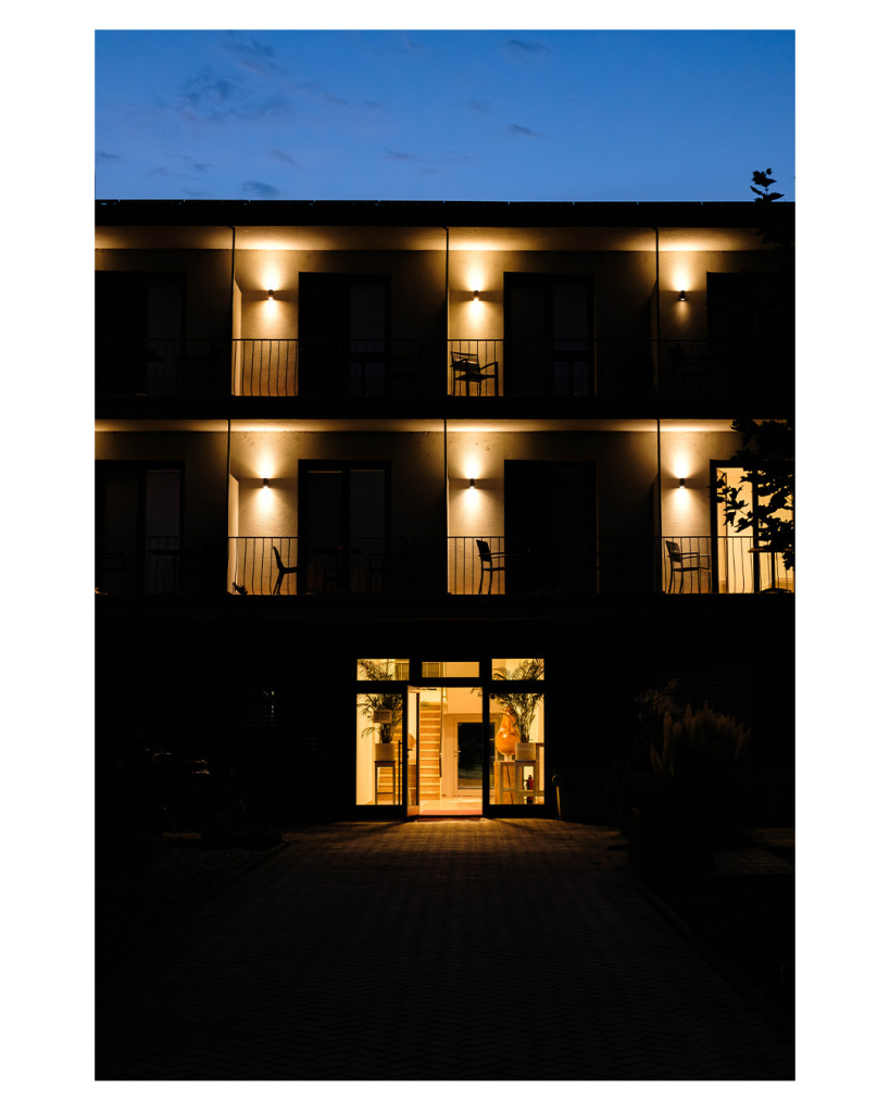 Foto im Hochformat. Nachts. Blick auf die Front einer Hotelanlage. Unten mittig ist eine offene Glastüre. Dahinter sieht man eine Treppe hoch gehen. Im ersten und zweiten Stock sind jeweils Balkons mit Geländer und Türen zu den Hotelräumen. Davor steht jeweils ein Stuhl. An jeder Türe ist ein Licht, welches die Türen spärlich beleuchtet. Der Großteil des Bildes versinkt im Dunkeln.