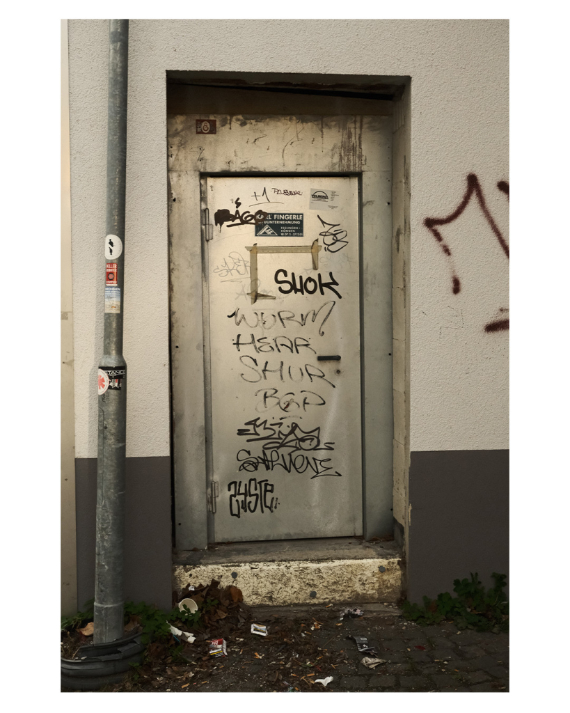 Foto im Hochformat. In einer weißen Hauswand ist eine Tür aus Stahl. Darauf sind Graffiti Tags mit schwarzem Stift geschrieben. Links neben der Tür ist ein Laternenpfahl auf dem verschiedene Aufkleber kleben. 