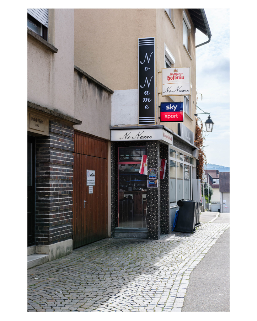 Foto im Hochformat. Blick auf ein Eckhaus in einer Stadt. In der Ecke ist die offene Türe zu einer Kneipe. Die Kneipe heißt No Name. Dies steht über der Tür, weiter oben auf einem schwarzen Schild und daneben auf einem weißen Schild. Es hängt außerdem ein Sky Sport Schild und eine VfB Flagge. Die Türe ist offen, drin ist eine Bar mit leeren Hockern. 