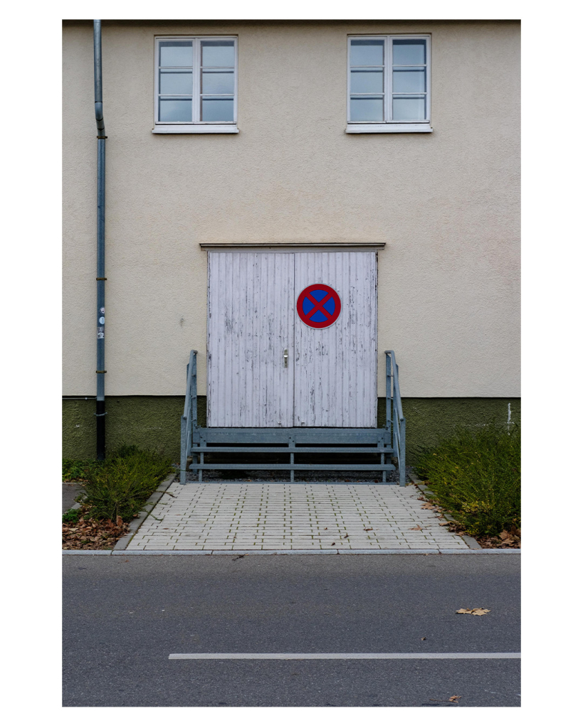 Foto im Hochformat. Blick über ein Straße auf ein Haus. Helle Hauswand. Mittig ist eine Doppeltür aus weiß angestrichenem alten Holz. Daran hängt ein absolutes Halteverbot Schild- Zur Tür führen drei Stufen eine Gittertreppe. 