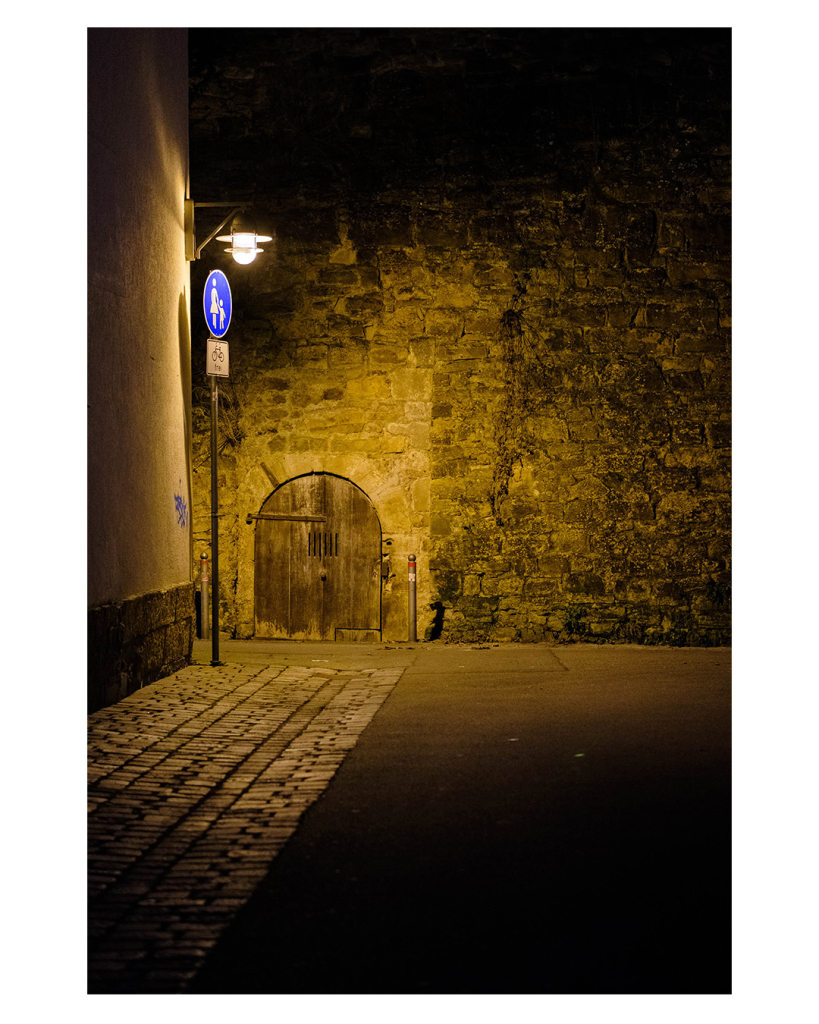 Foto im Hochformat. Nachts. Altes Mauerwerk aus einiger Entfernung. Links ist eine alte hölzerne Tür mit Torbogen. Davor, an einem anderen Haus, ist eine Straßenlaterne, die alles erleuchtet. Nach außen hin wird das Bild immer dunkler. 