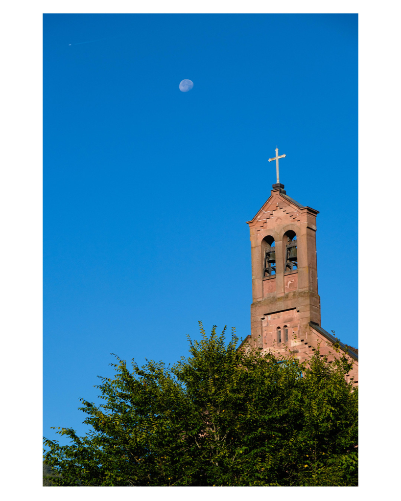 Foto im Hochformat. Blick Richtung blauem Himmel. Unten ragt die Krone eines Baumes ins Bild, dahinter ist der flache Turm einer Kirche. Oben sind zwei Glocken, darüber ein Kreuz. Am Himmel ist der Mond. 